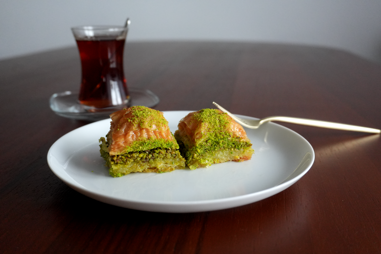 Baklava mit Pistazienfüllung auf Teller, feiner Yufka-Teig, pistaziengrüner Belag, Tee im Hintergrund